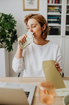 Smiling woman with closed eyes holding money at desk with envelope and laptop, feeling content.